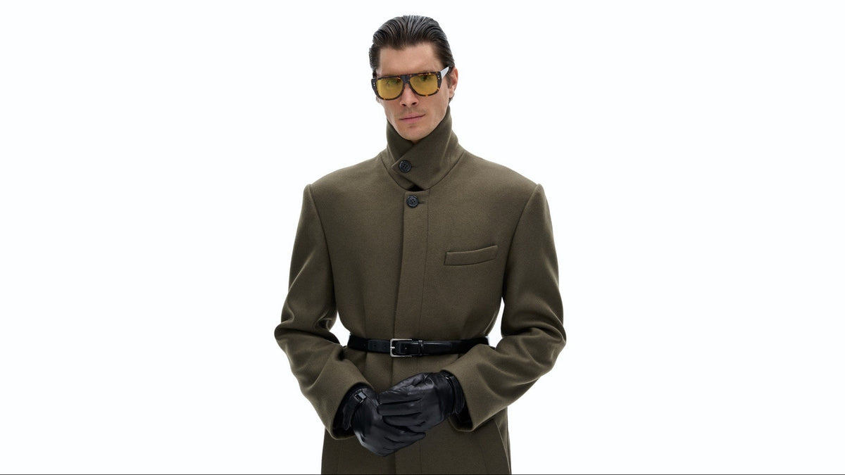 A man wearing a dark olive green military-style coat with a high collar, black belt, black gloves, and yellow-tinted sunglasses, standing against a solid white background.